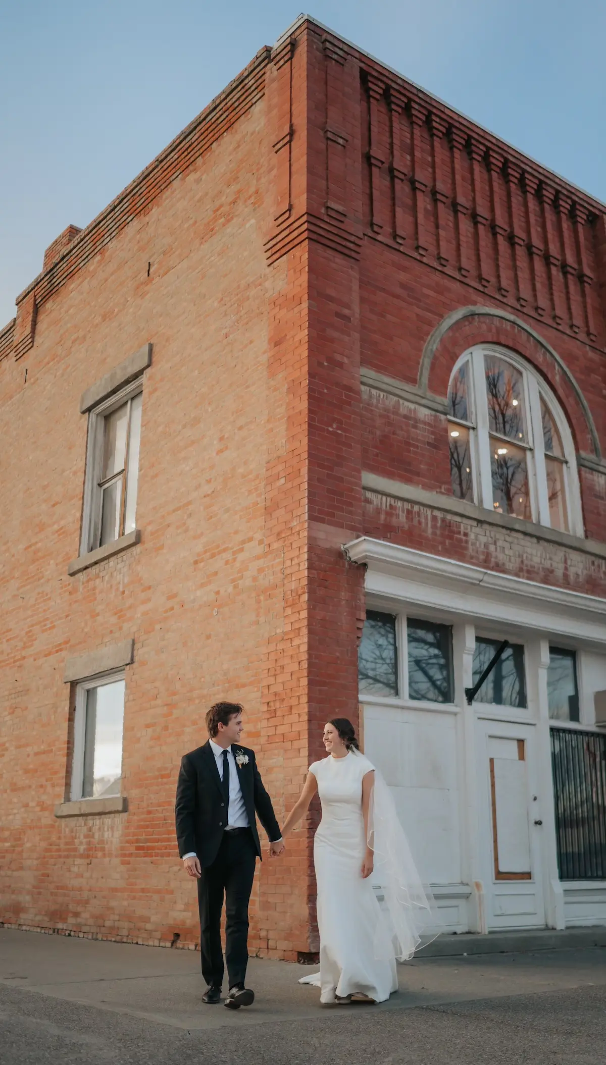 The couple standing hand in hand outside the historic brick wedding venue with a blue sky above