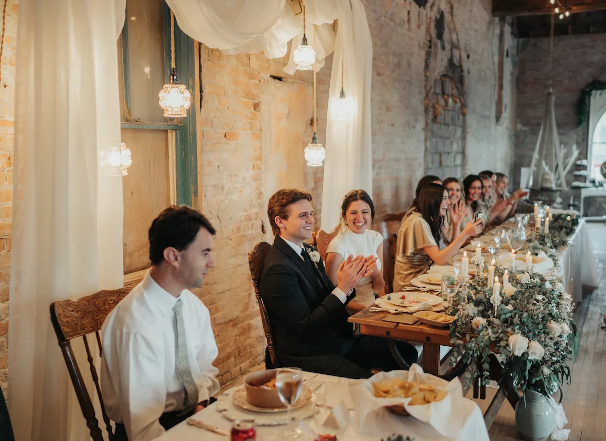The couple laughing at the sweetheart table during dinner, the room glowing with candlelight and vintage lanterns.