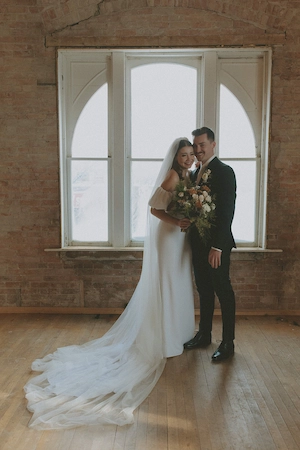Bride in a long white gown and veil holding a bouquet, standing and smiling with groom in a black suit in front of a large arched window with brick walls.