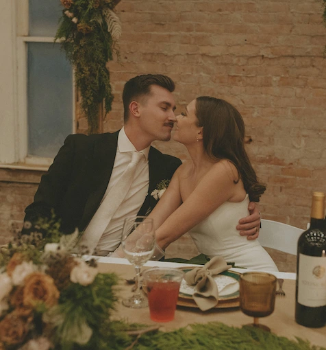 Bride and groom sitting at a decorated wedding table, leaning in for a kiss with a brick wall background.