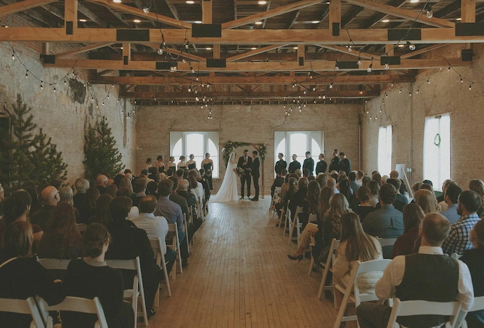 Indoor wedding ceremony with bride and groom standing under a wooden arch in front of seated guests in a rustic venue with exposed brick walls and wooden beams.