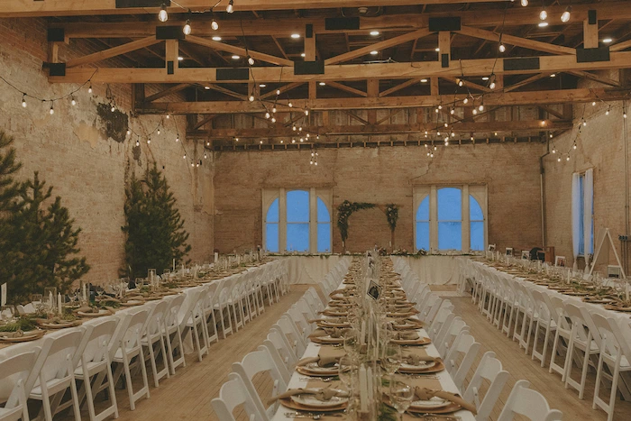 Rustic banquet setup in a brick loft hall with long tables set for a formal dinner, white chairs, string lights, and pine trees along the walls.