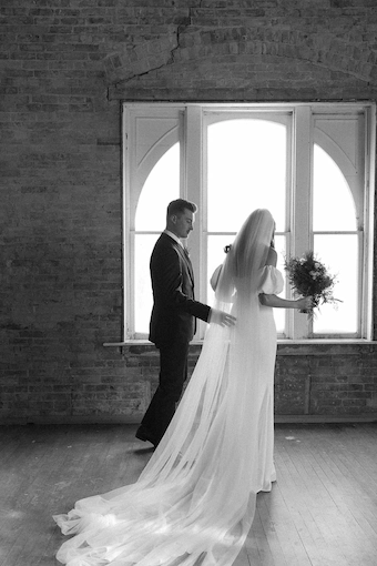 Black and white photo of a bride and groom standing by a large arched window with the bride holding a bouquet and wearing a long veil and gown.