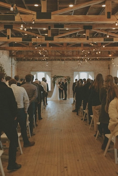 Bride and groom standing at the altar in a rustic venue with wood beams overhead with guests standing on both sides.