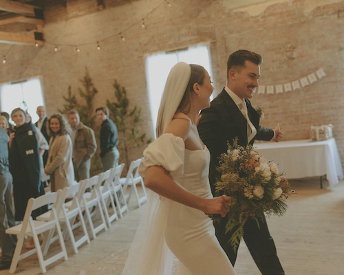 Bride and groom joyfully walking hand in hand inside a rustic wedding venue with guests seated in white chairs.