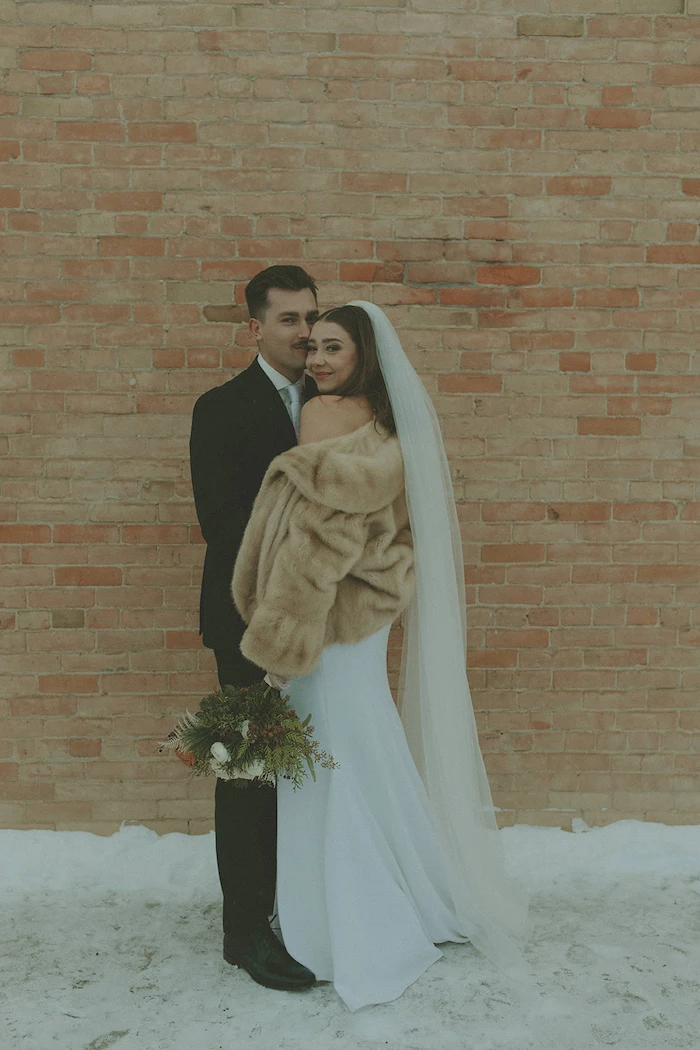Bride in a white dress and beige fur shawl holding a bouquet stands with groom in a black suit against a brick wall on snow-covered ground.