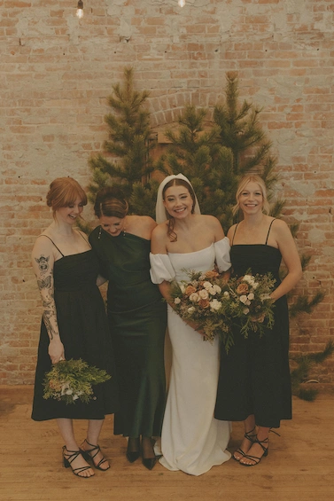 Bride in white dress and three bridesmaids in black dresses standing in front of pine trees and a brick wall, holding bouquets and smiling.