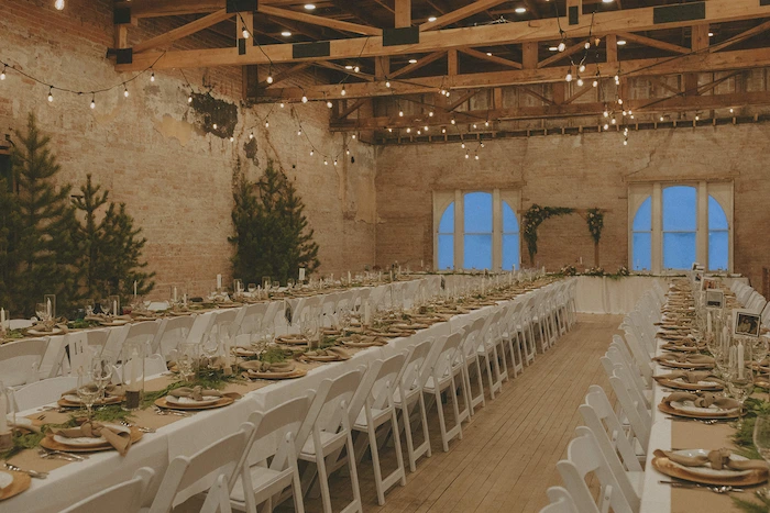 Long banquet tables with white chairs set for a rustic indoor event in a wooden-beamed room decorated with string lights and pine trees.