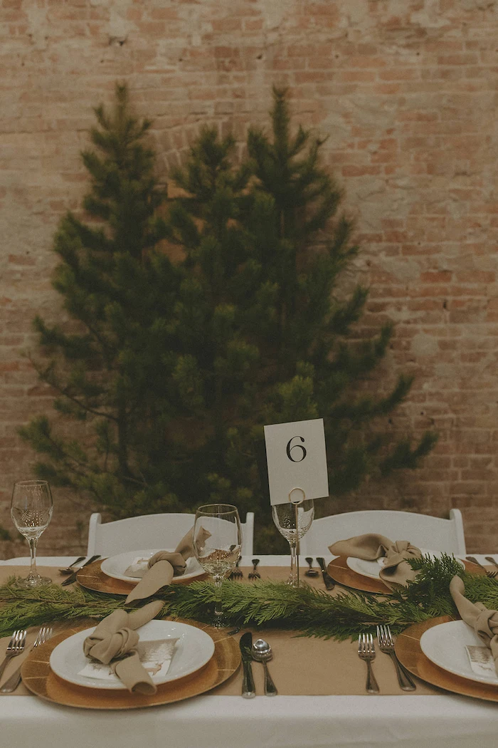 Elegant table setting with white plates, beige napkins, wine glasses, and green foliage centerpiece against a brick wall with a pine tree backdrop.