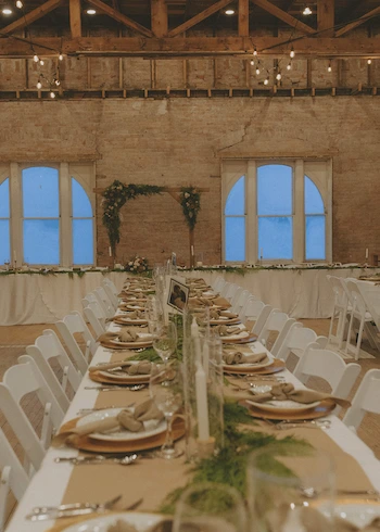 Long banquet table set with plates, napkins, glassware, and greenery in a rustic venue with brick walls and wooden beams.