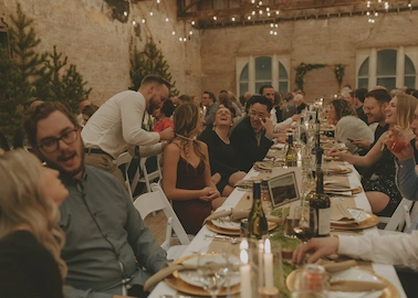 Group of people enjoying a festive dinner party at a long, candlelit table in a rustic, warmly lit room.