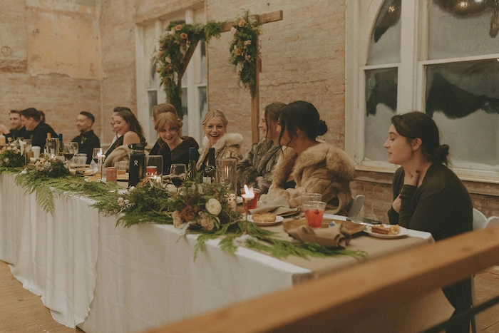 Group of people sitting at a long white-clothed table decorated with greenery and flowers, enjoying a wedding social event in a rustic indoor setting.