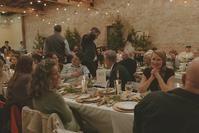 Guests seated at banquet tables enjoying a meal in a warmly lit rustic event space with string lights and brick walls.