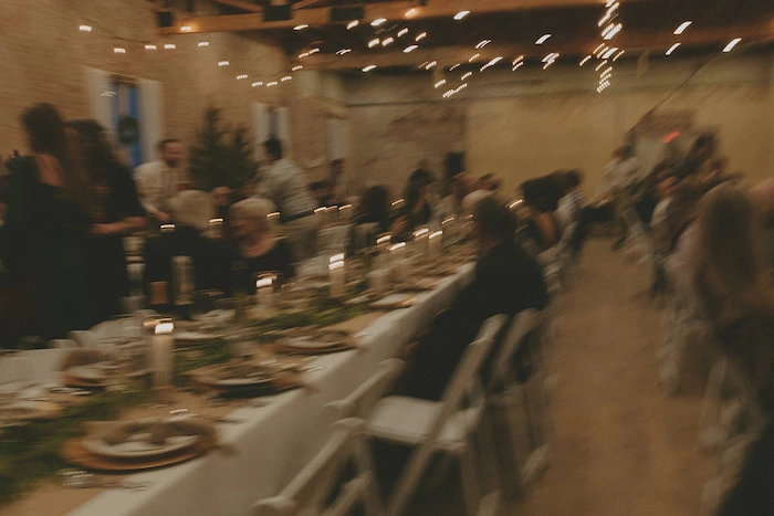 Blurry image of people seated and standing around a long candlelit dinner table in a rustic indoor setting with string lights above.