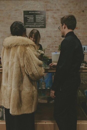 Two people standing at a counter with a woman behind it in a brick-walled space, one person wearing a light brown fur coat and the other in black clothing with glasses.