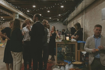 People socializing indoors at a dimly lit event space with string lights overhead and a bar with snacks and drinks.