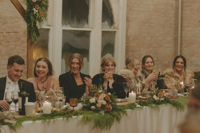 A group of six people from the bridal party sitting at a decorated head table with flowers and candles, smiling and clapping, at a wedding banquet.