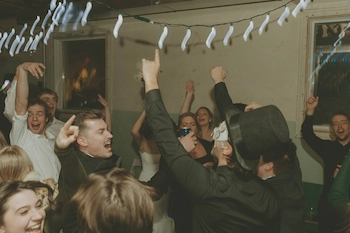 Group of people dancing and celebrating indoors under string lights with joyful expressions.