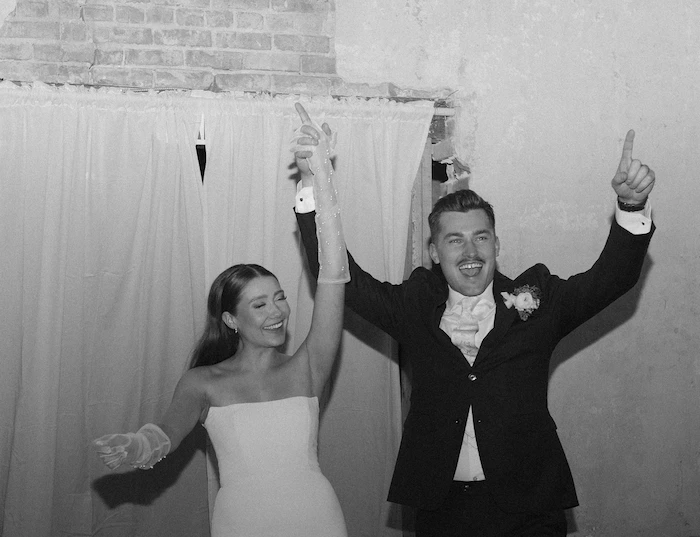 Black and white image of Bride and groom celebrating joyfully with raised hands in front of a brick wall and curtains.