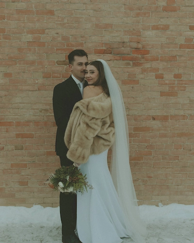 Bride in white dress and fur shawl with long veil holding bouquet embraces groom in black suit in front of brick wall with snow on ground.