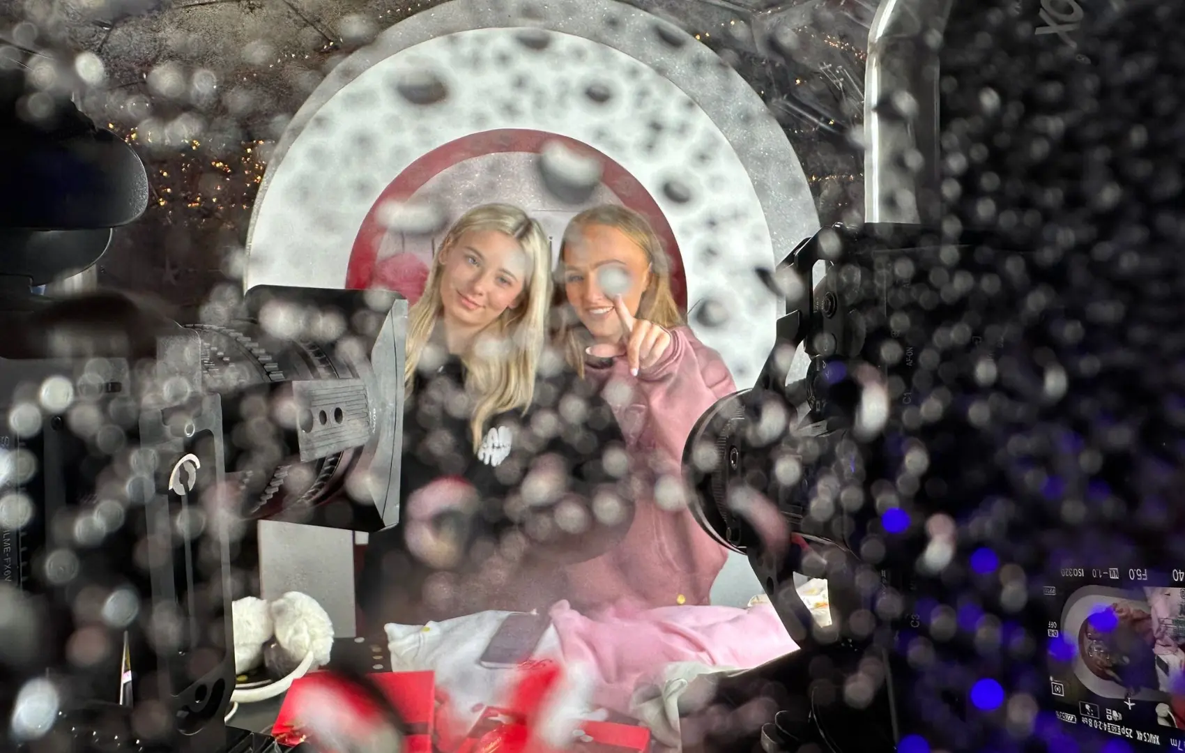 Two women are standing in front of a camera with a red circle on it.