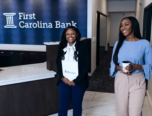 Two smiling women standing inside a First Carolina Bank office, one holding a branded folder, the other a coffee cup.