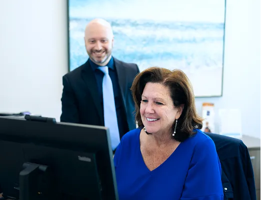 Smiling woman in blue blouse working on a computer with a man in a suit standing behind her.