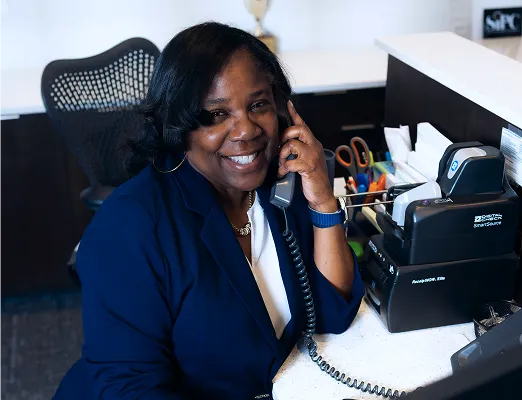 Smiling woman in blue blazer talking on the phone at an office desk with stationery and a printer.