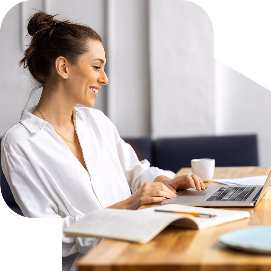 Woman smiling while typing on a laptop at a wooden table with a notebook, pen, and coffee cup.