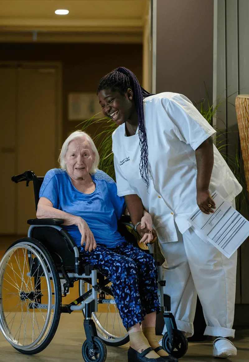 Une aide-soignante accompagne une résidente en fauteuil roulant dans un établissement