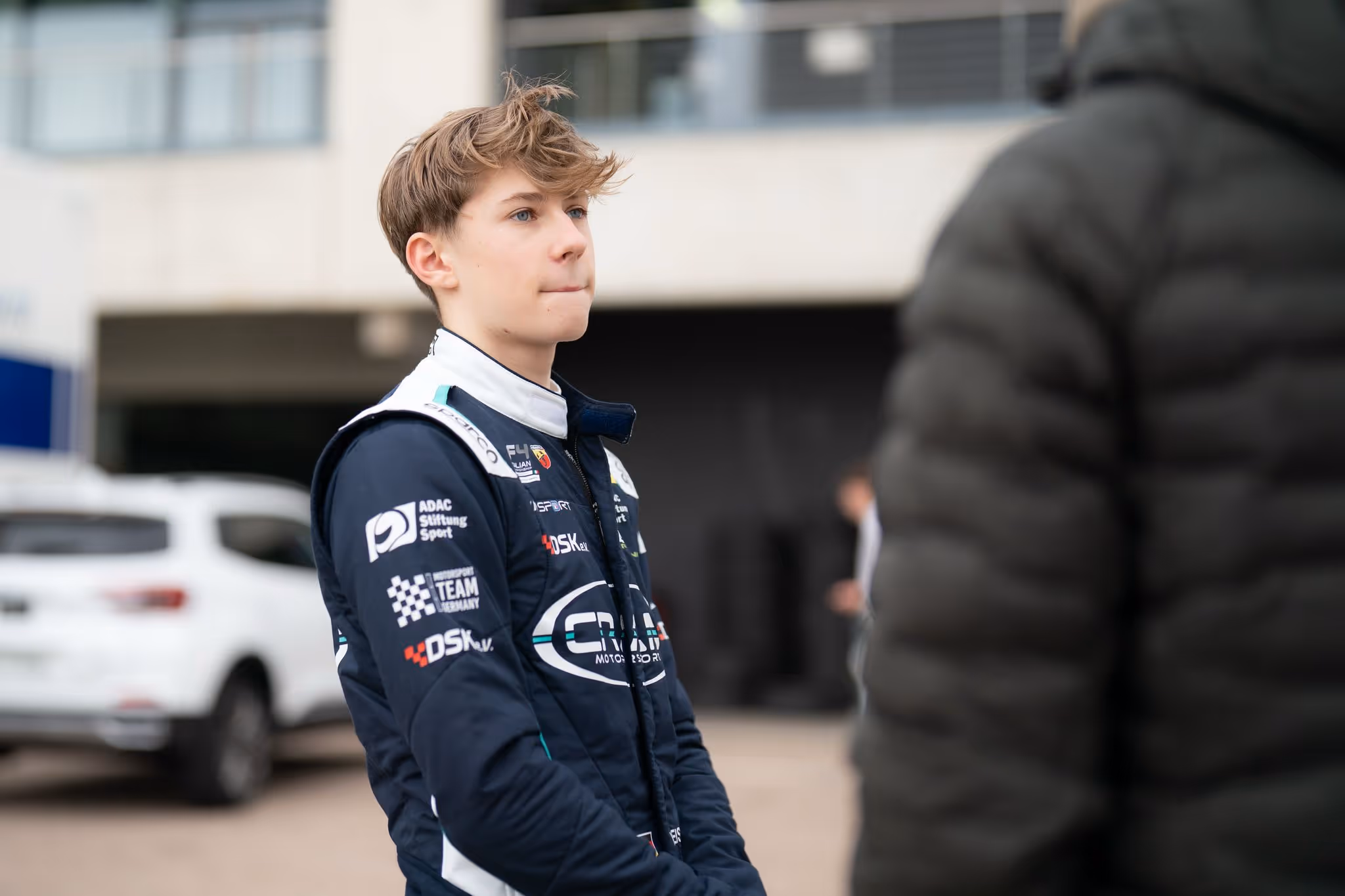 Young male race car driver in blue racing suit with sponsor logos stands outside near a white vehicle, looking ahead.