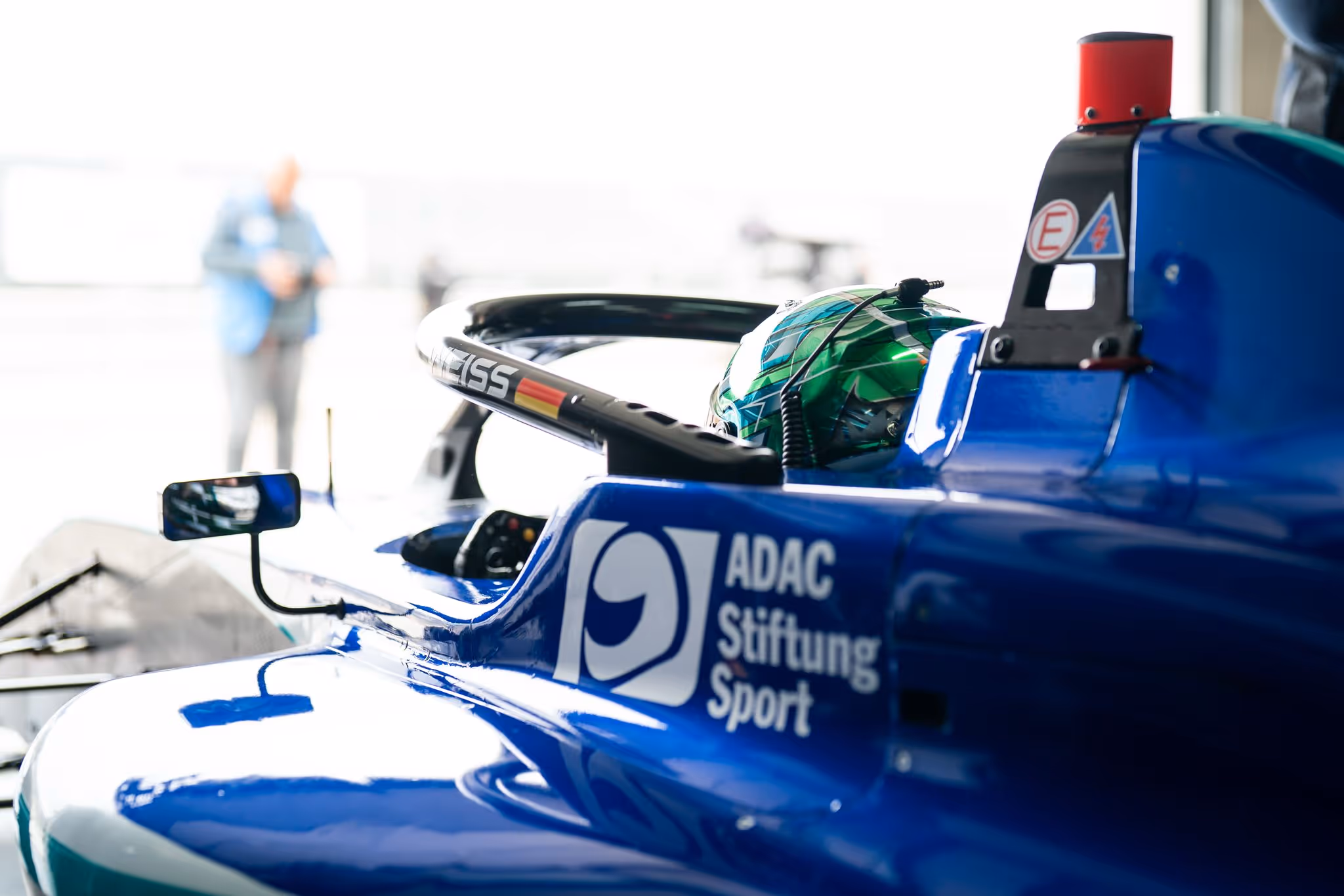 Close-up of a race car driver in a blue formula car with a green helmet and ADAC Stiftung Sport logo.