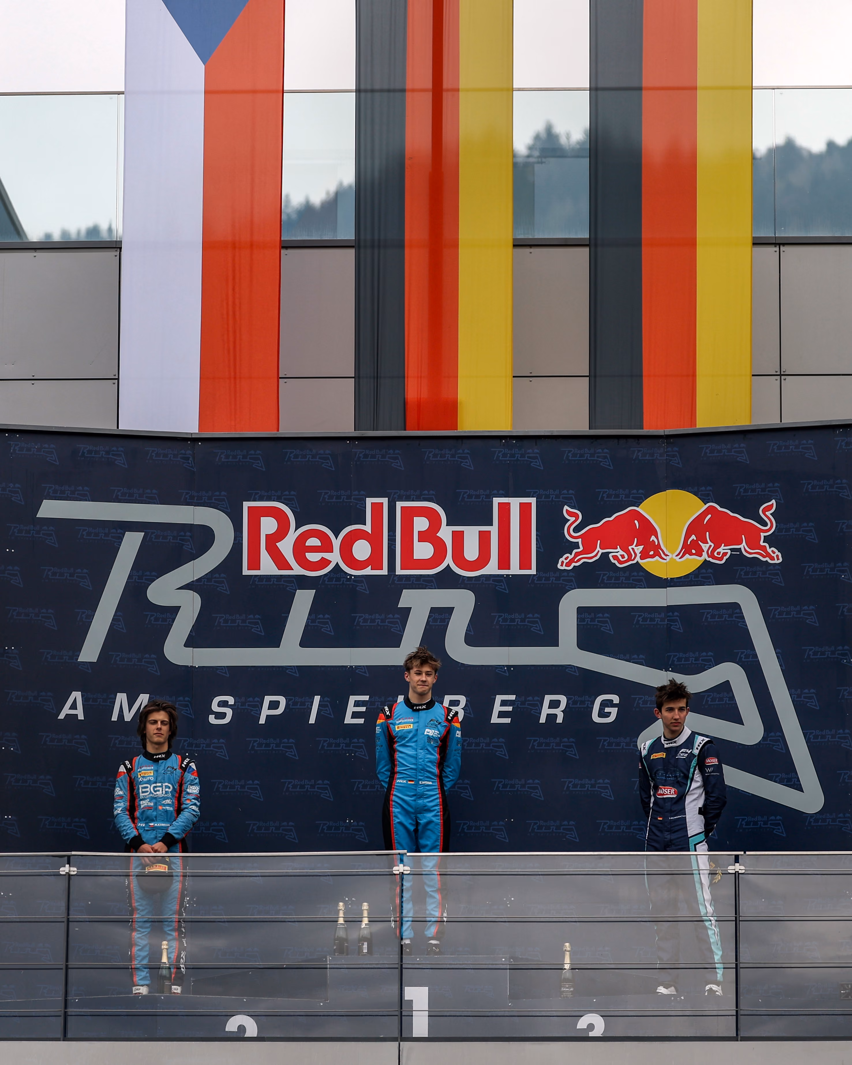 Elia Weiss standing on the Red Bull Ring podium at Spielberg, with flags above and champagne bottles in front.