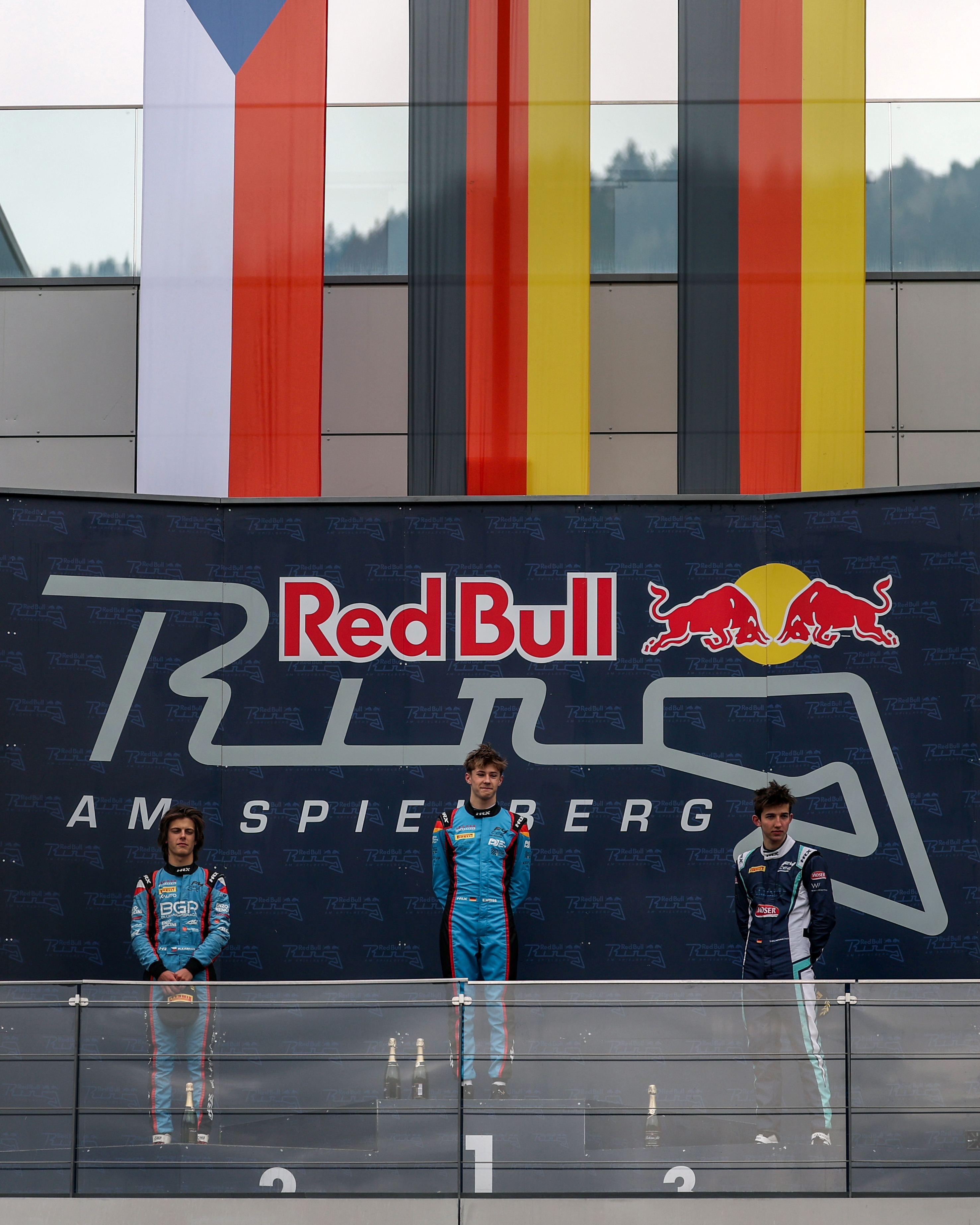 Elia Weiss standing on the Red Bull Ring podium at Spielberg, with flags above and champagne bottles in front.