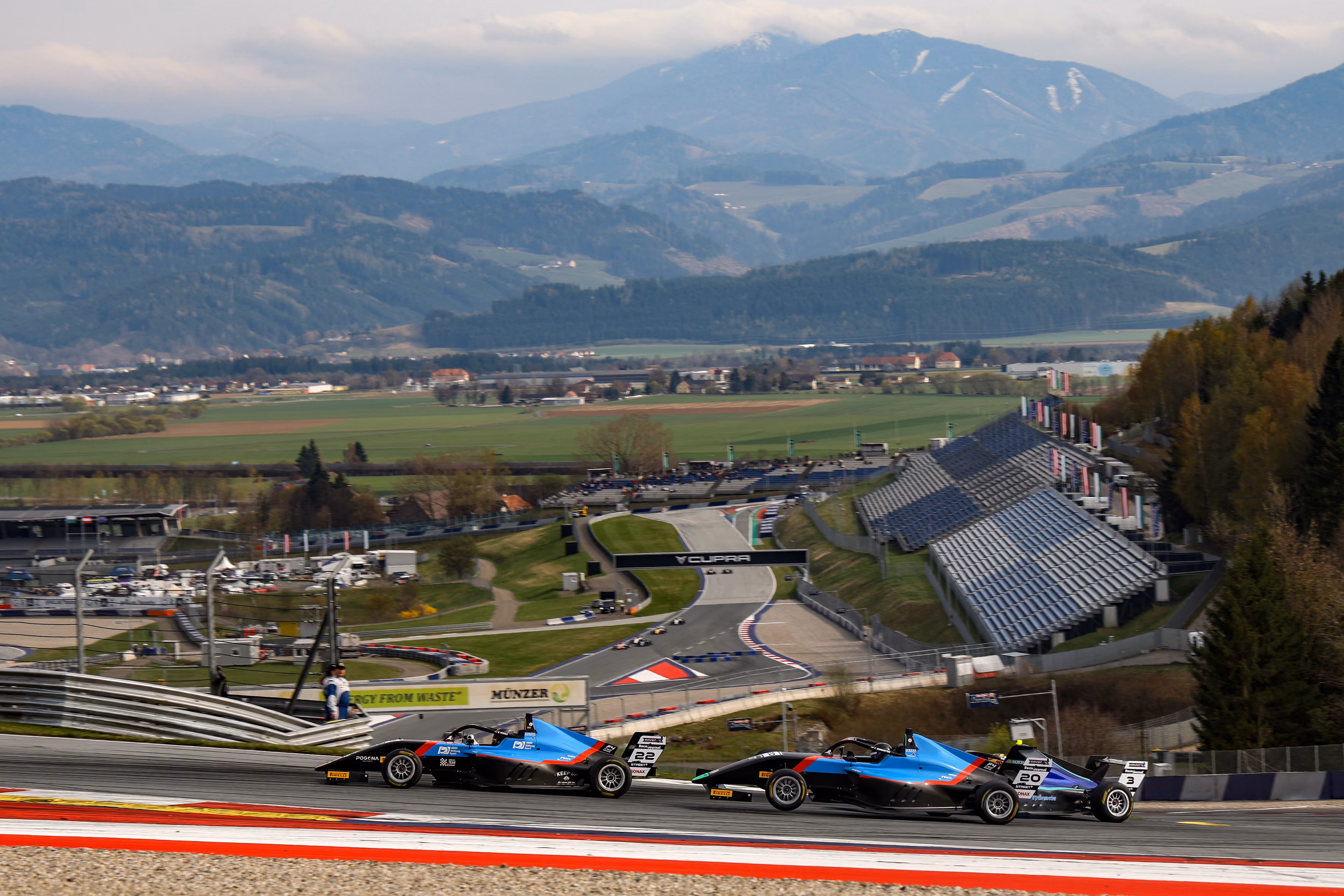 Elia Weiss in his Formular 4 car on a curved section of a racetrack with grandstands and mountainous landscape in the background.