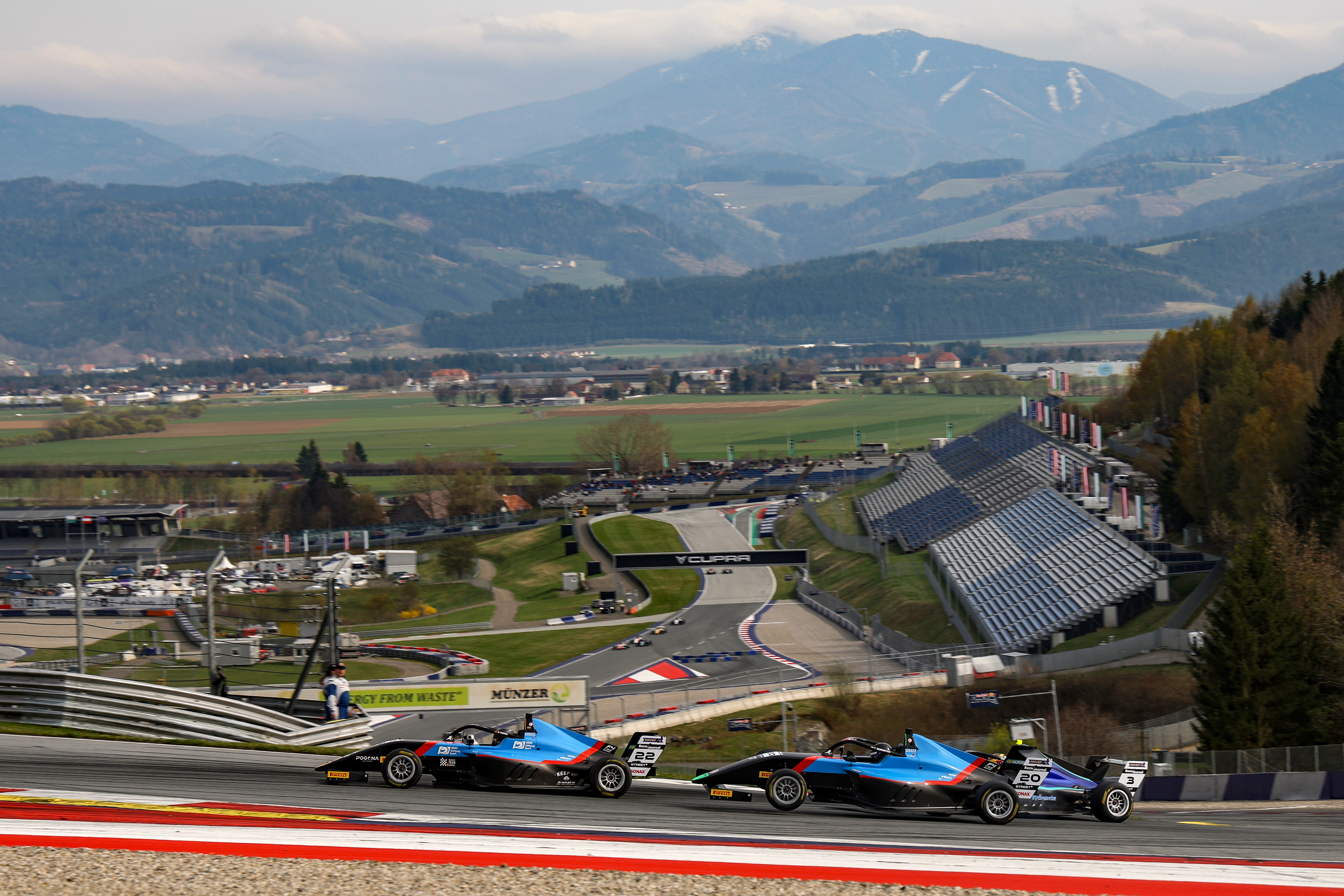 Elia Weiss in his Formular 4 car on a curved section of a racetrack with grandstands and mountainous landscape in the background.