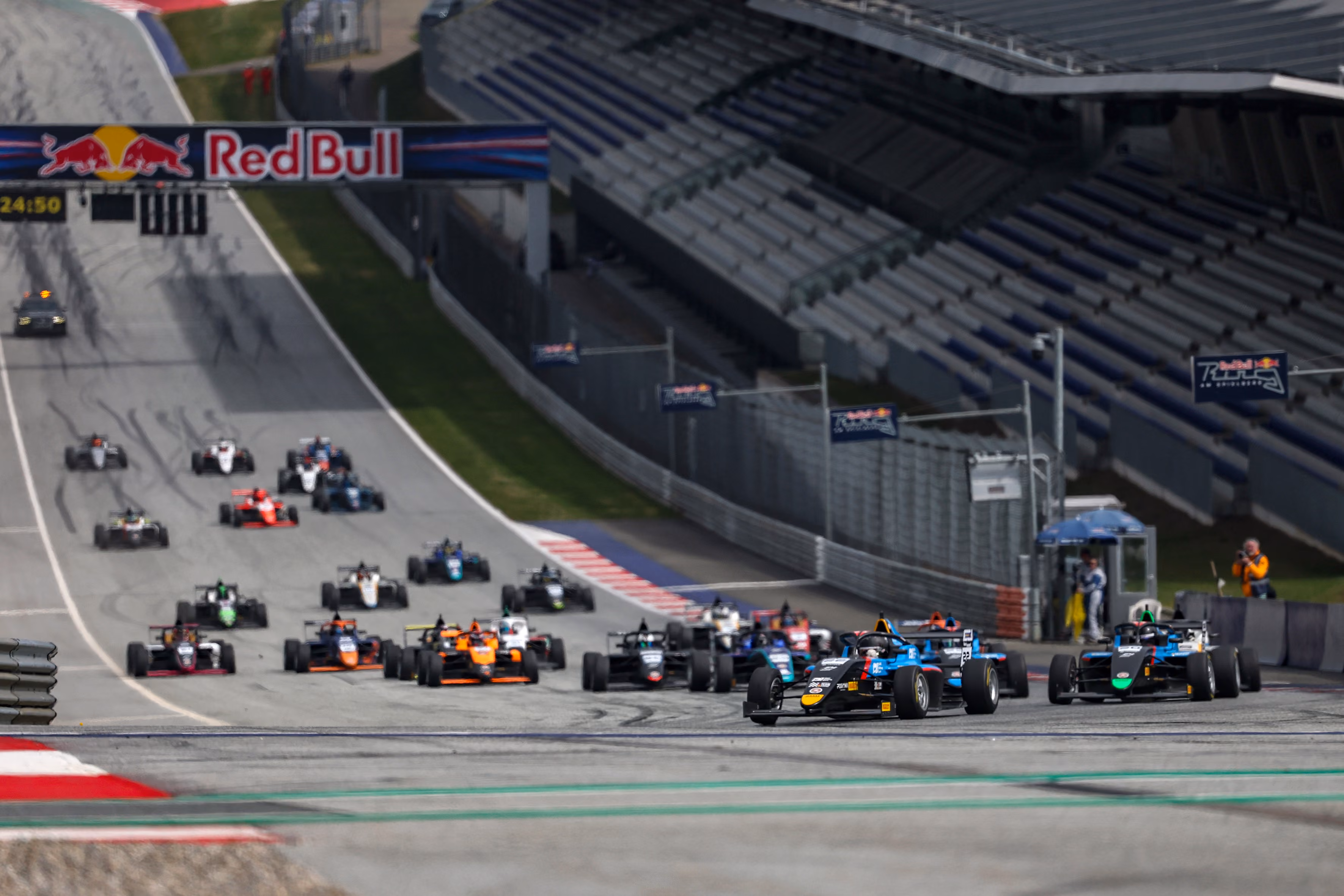 F4 race cars speeding on a track at the Red Bull Ring with grandstands in the background.