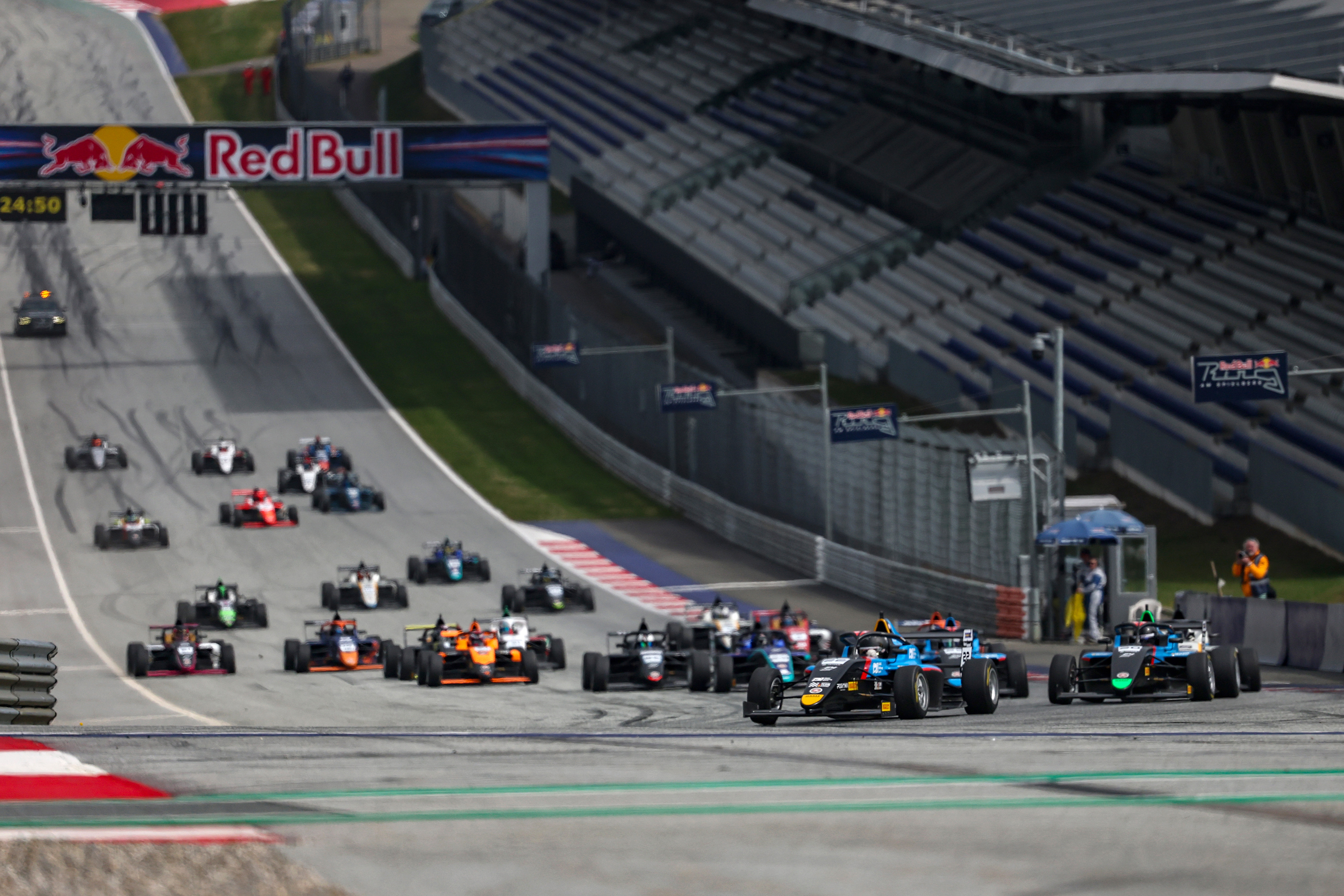 F4 race cars speeding on a track at the Red Bull Ring with grandstands in the background.