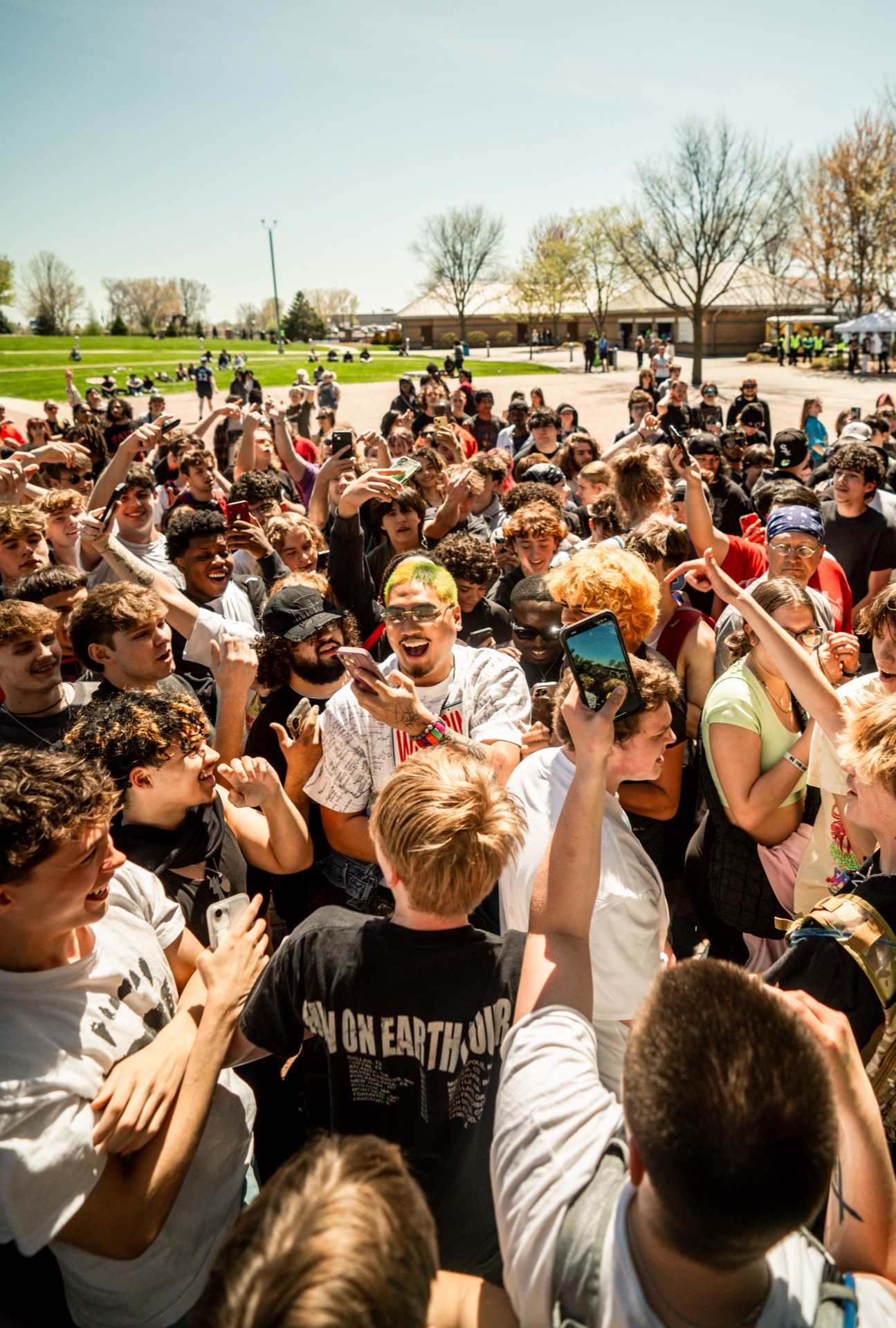 Dense crowd of cheering young people outdoors filming on phones at a daytime gathering