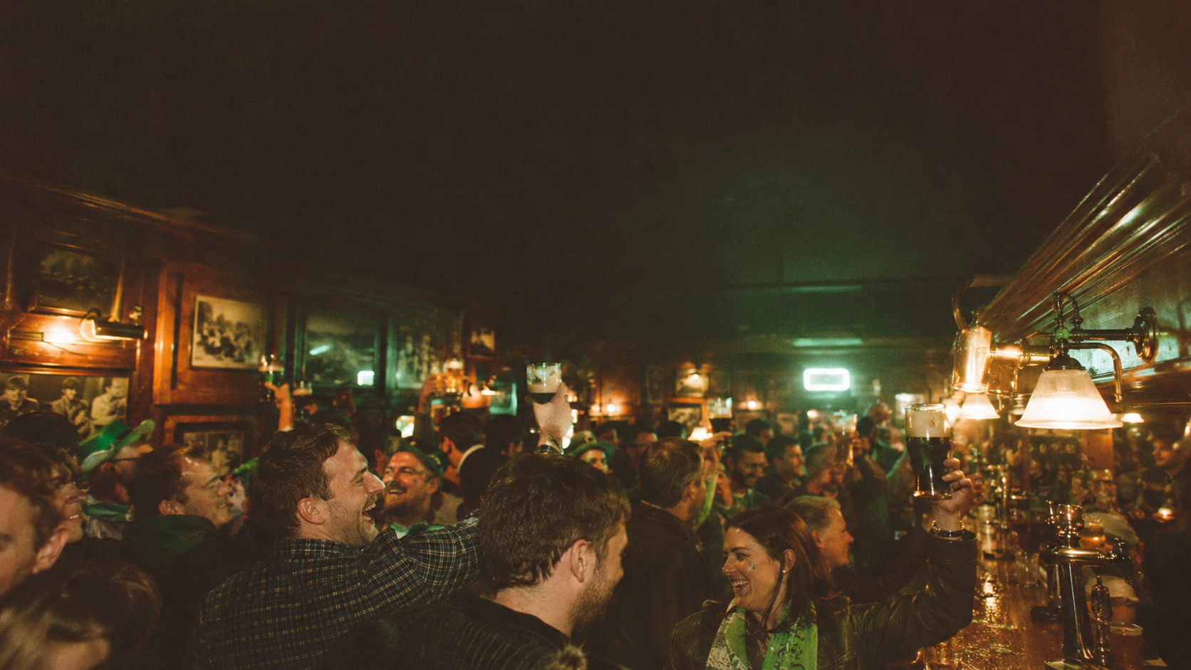 Packed dim pub crowd celebrating under green lighting, some in St. Patrick's Day attire