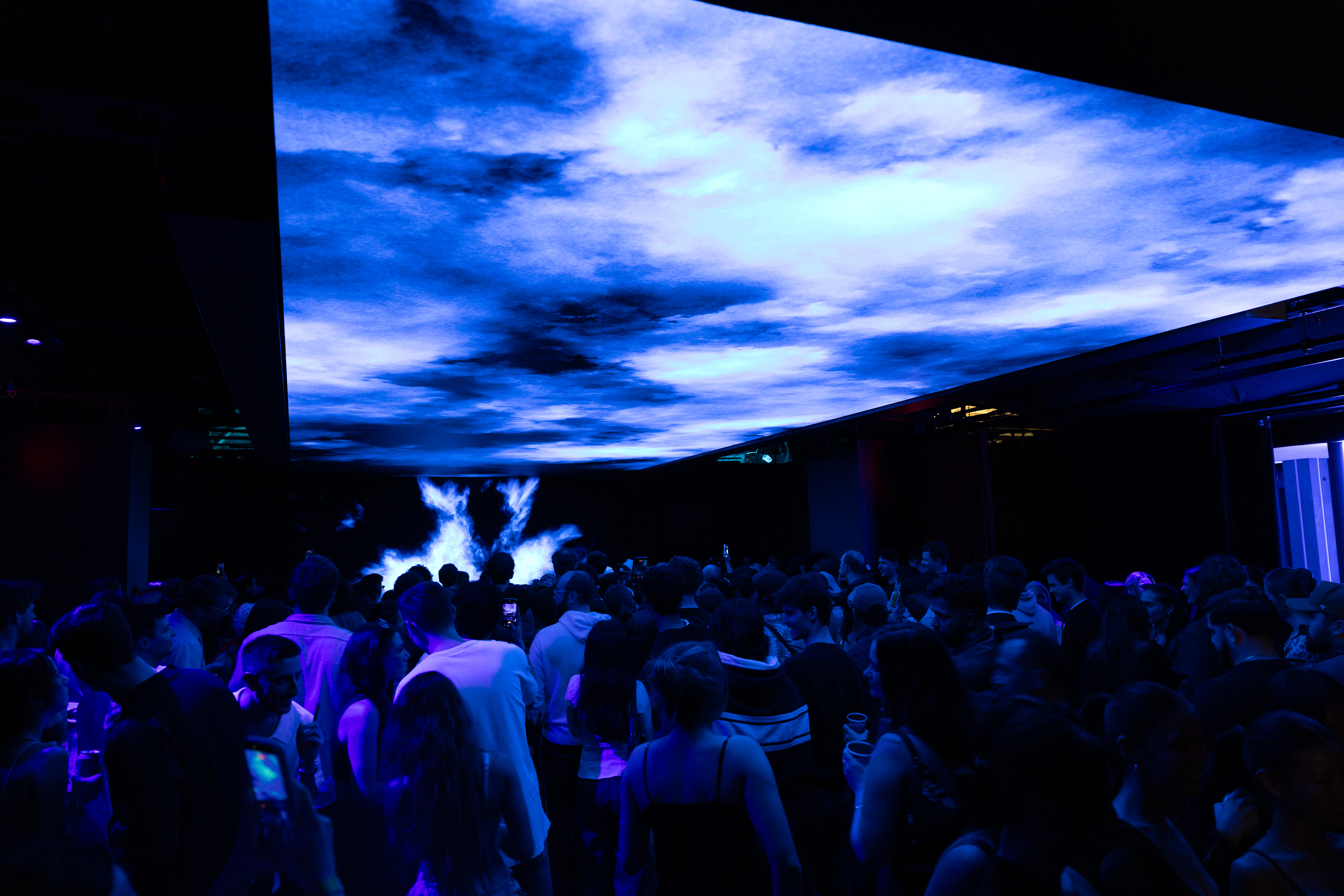 Crowd at a venue beneath a huge screen projecting blue sky and clouds