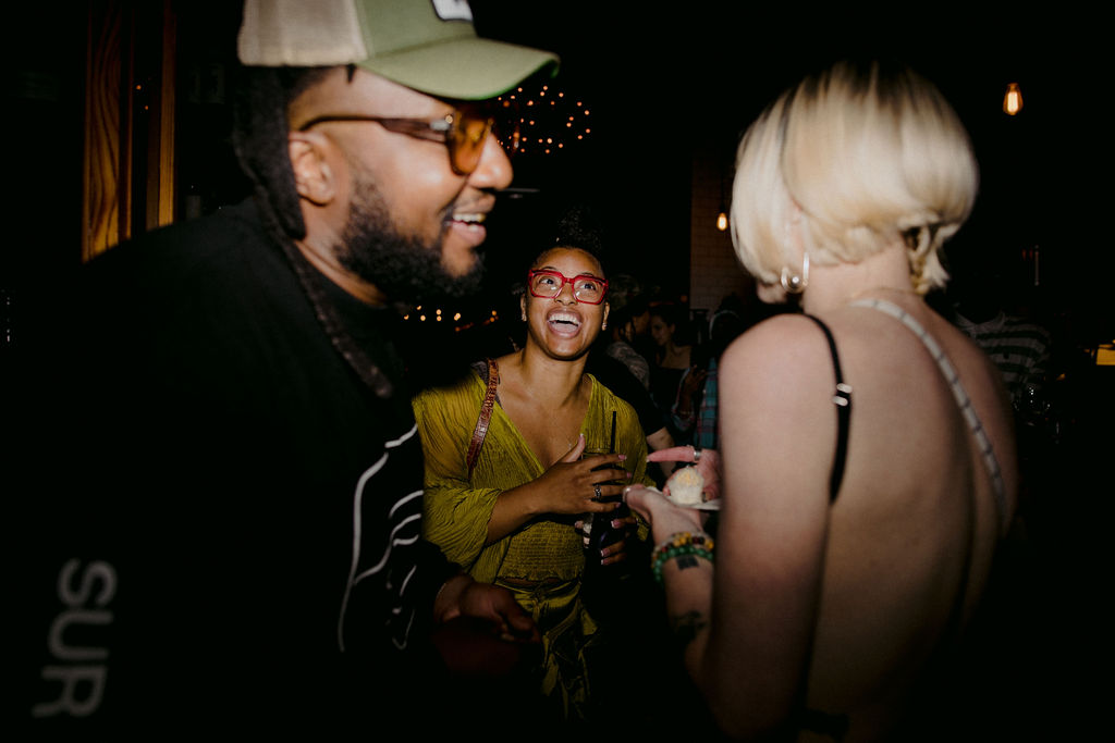 Three friends laughing and chatting over drinks at a dark, string-lit bar