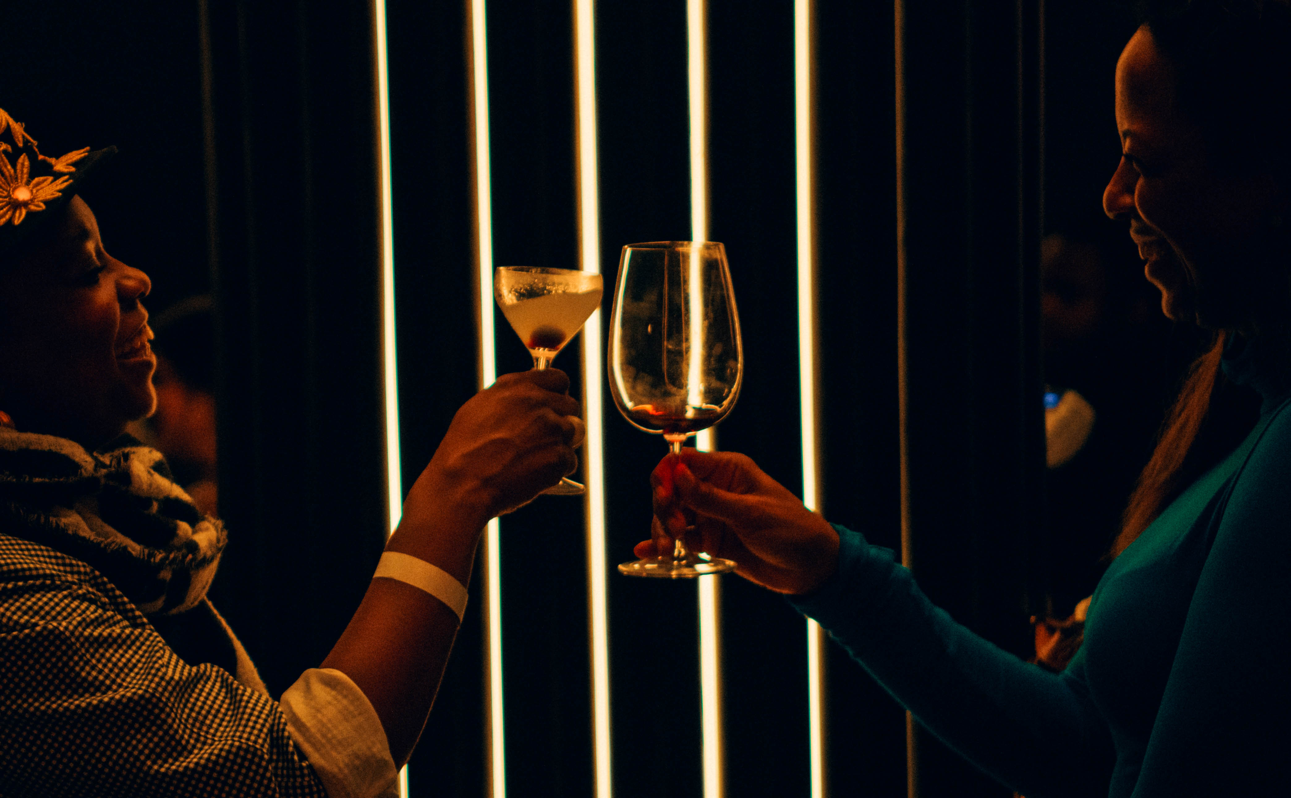 Two women toast a cocktail and wine glass before glowing vertical light bars