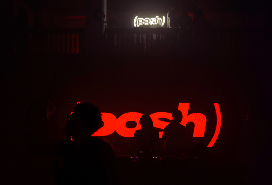 Glowing red 'posh' logo projection behind a silhouetted figure in a dark venue