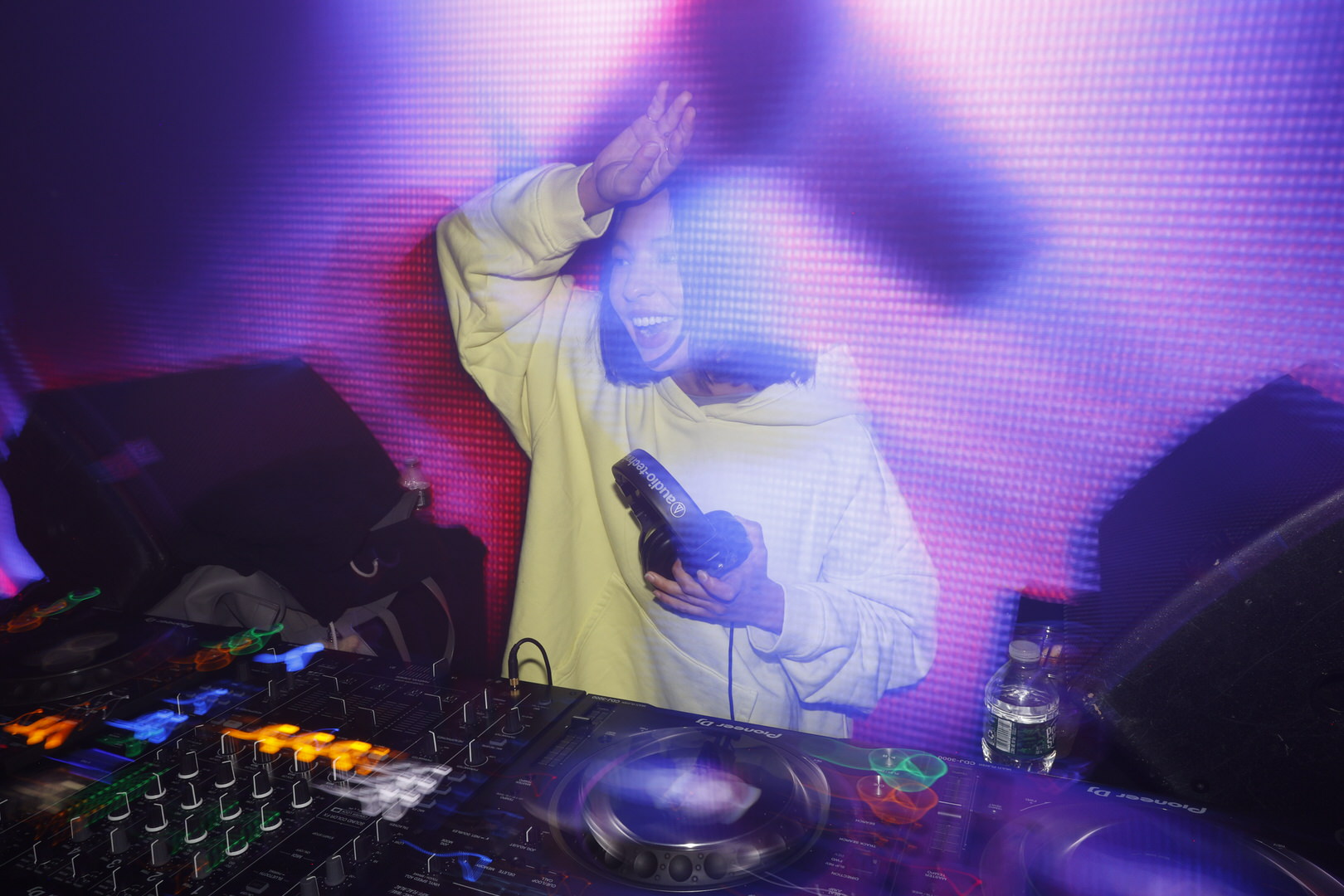 DJ in a white hoodie raising one arm, holding headphones over a glowing Pioneer mixer