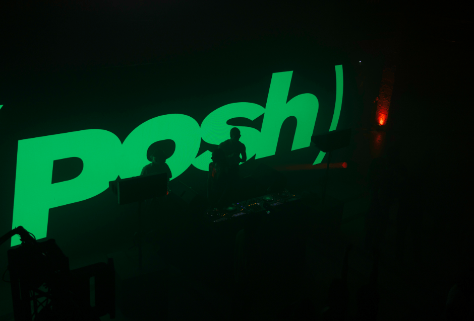 DJ performing behind a large glowing green 'Posh' logo on stage