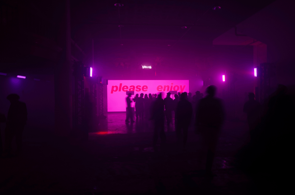 Silhouetted crowd before a pink 'please enjoy' sign in a dark hall