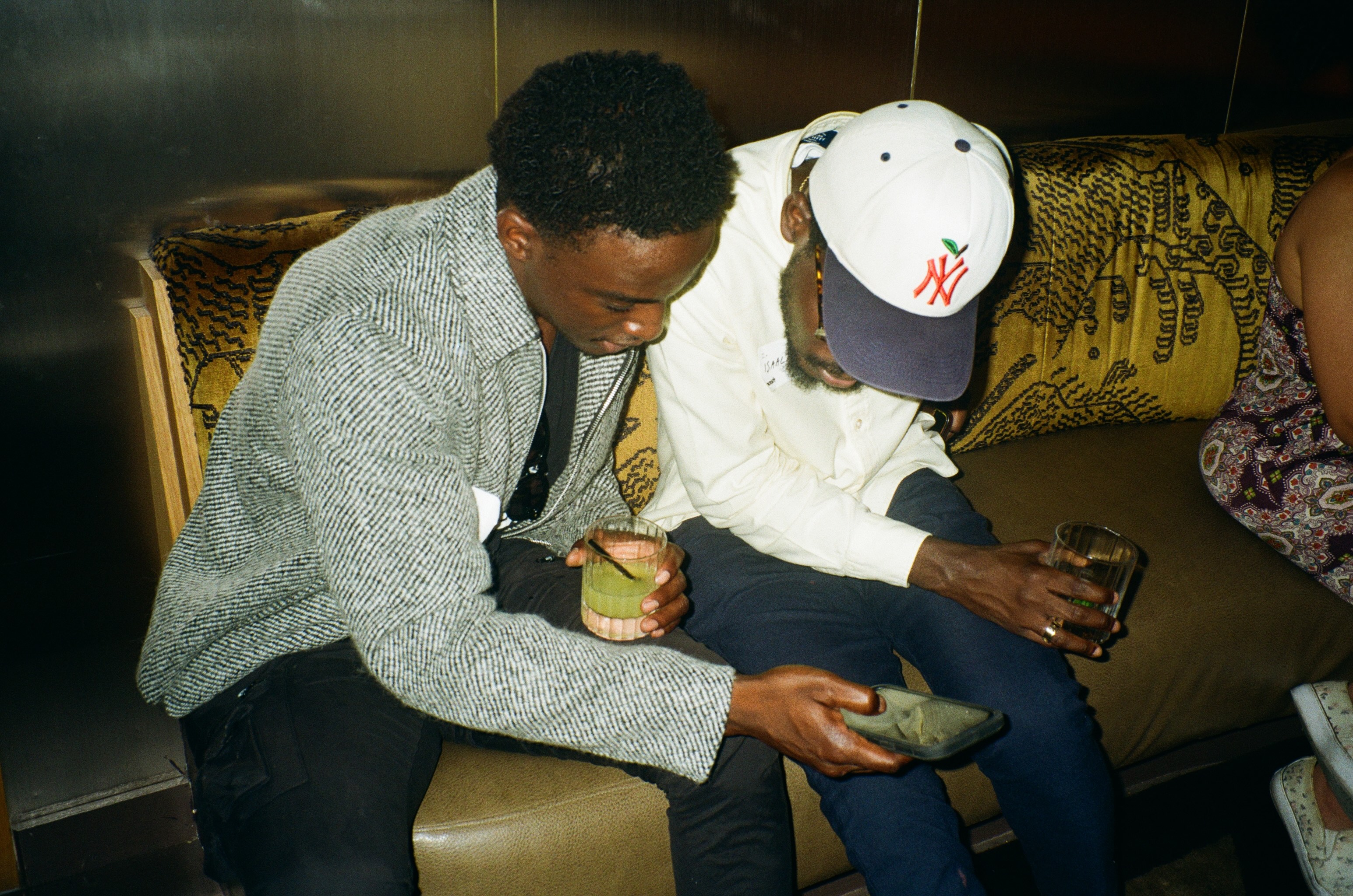 Two men on a club banquette, one sipping a cocktail, the other in a Yankees cap