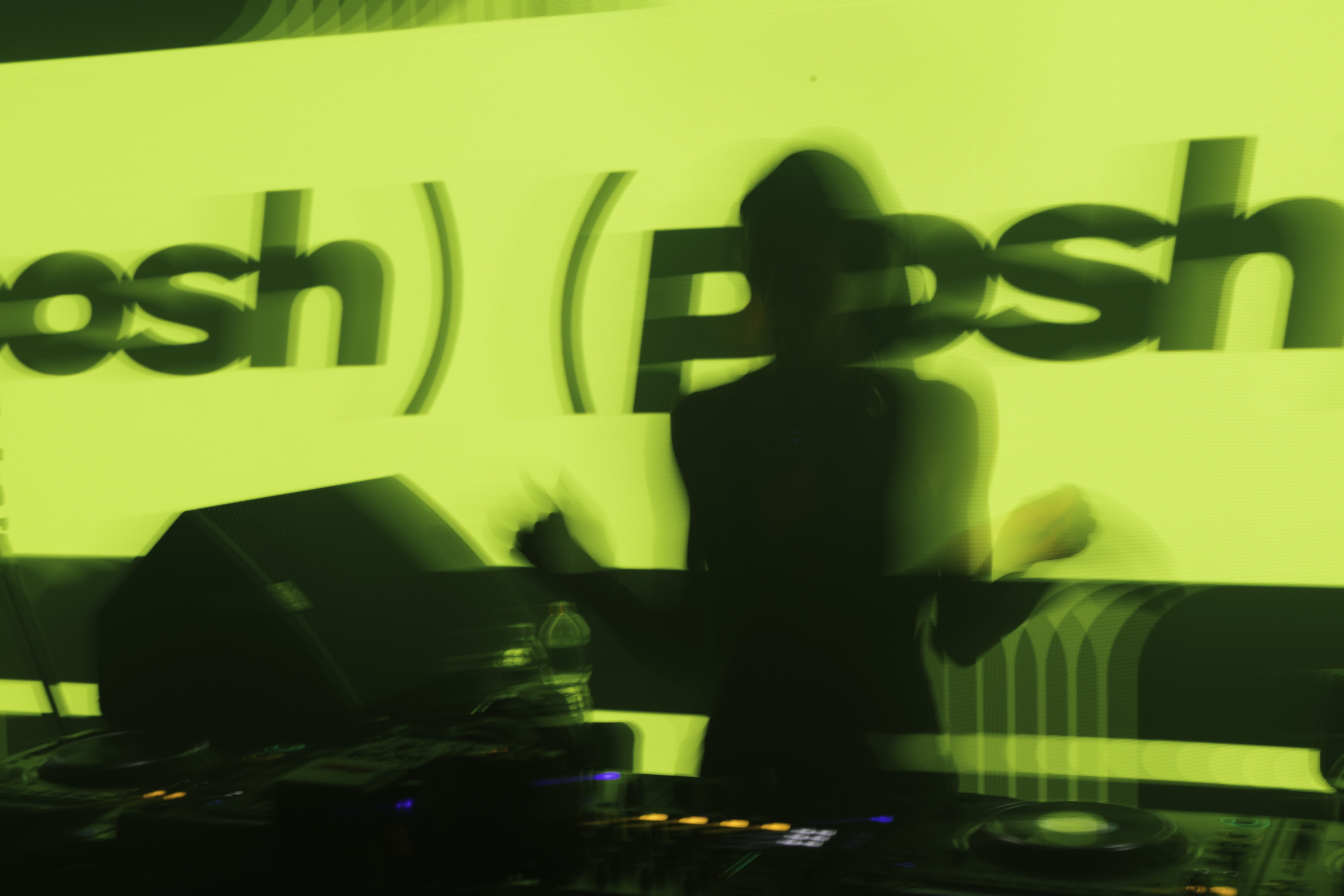 Silhouetted DJ at the decks against a glowing green wall projecting the Posh logo