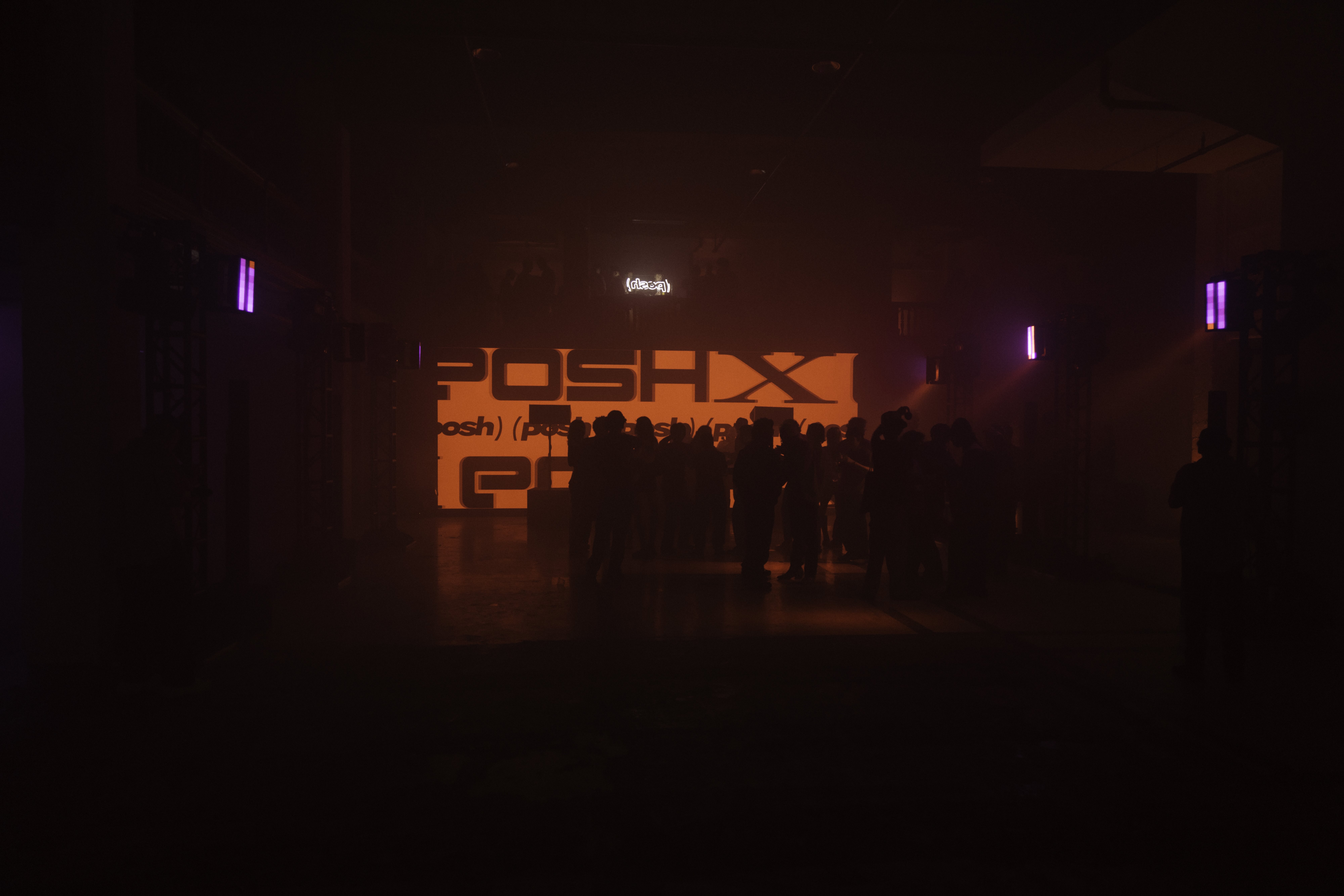 Crowd silhouetted before an illuminated orange Posh stage backdrop in a dark warehouse